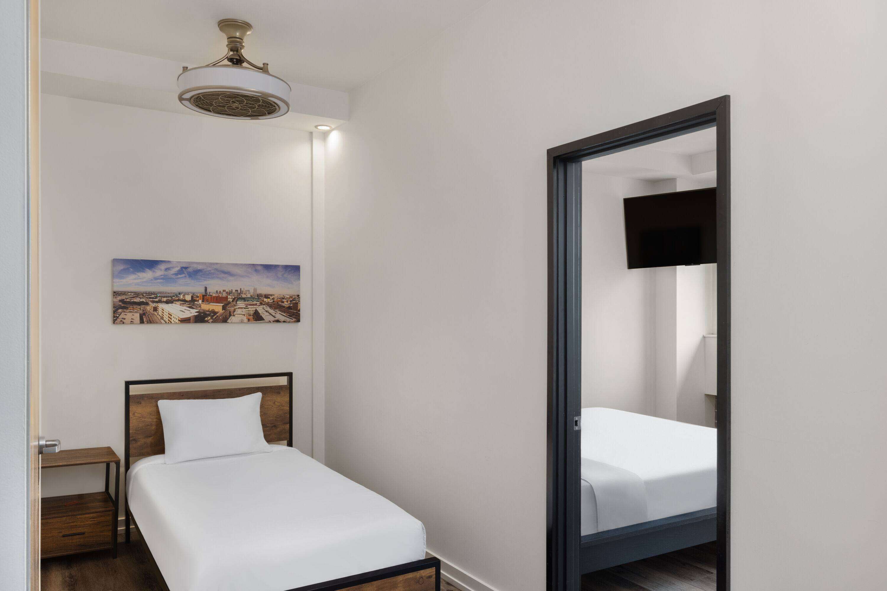 A hotel room with a bed, a nightstand, a painting on the wall, and a ceiling fan.