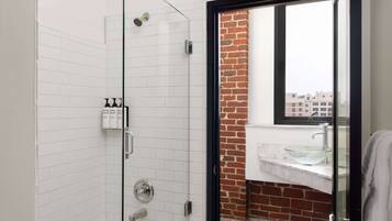 Un baño moderno con ducha de vidrio, paredes de azulejos blancos, una pared de ladrillo decorativa y vistas a edificios a través de una ventana.