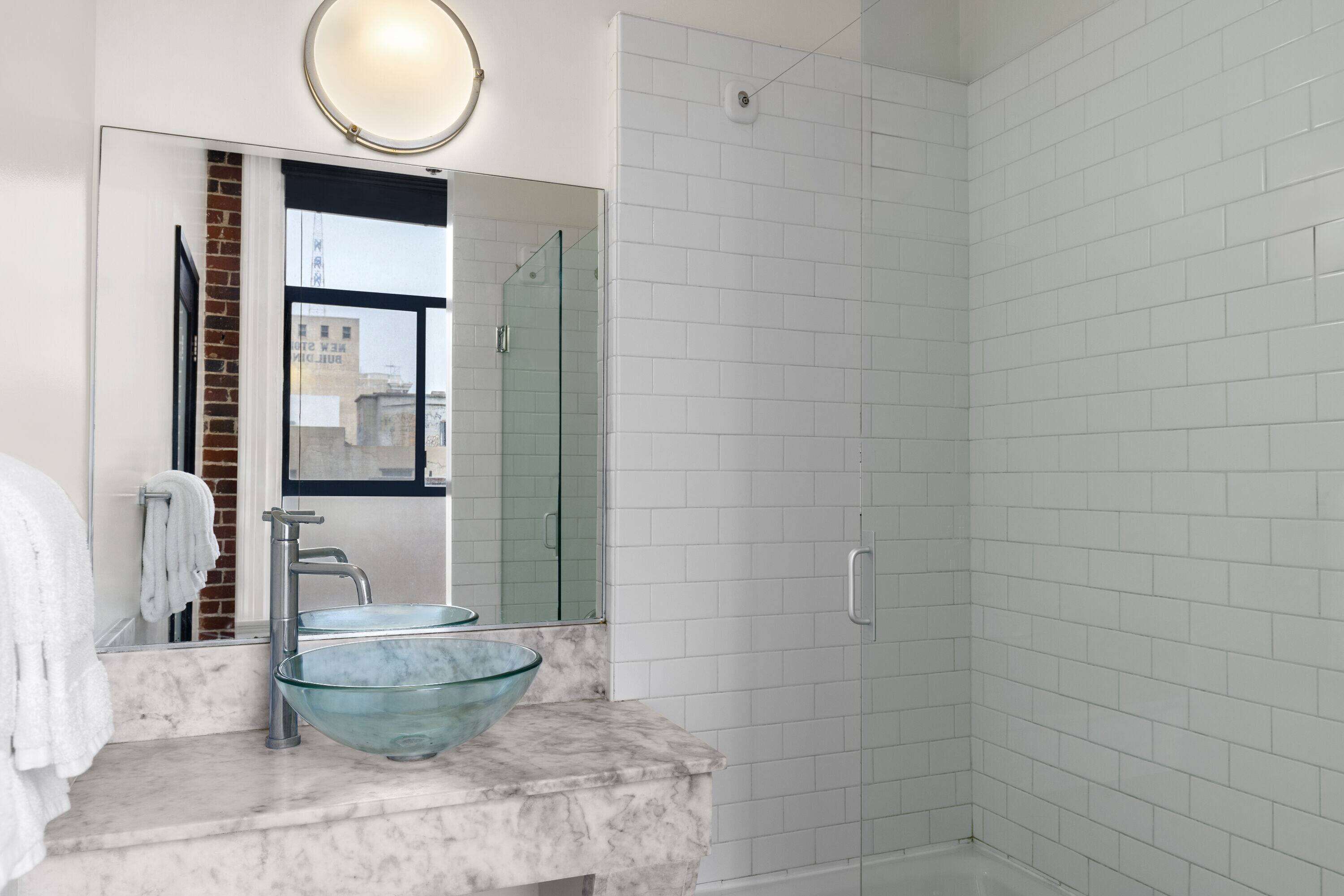 A bathroom with a glass bowl sink, a mirror, and a shower area with white tiles.