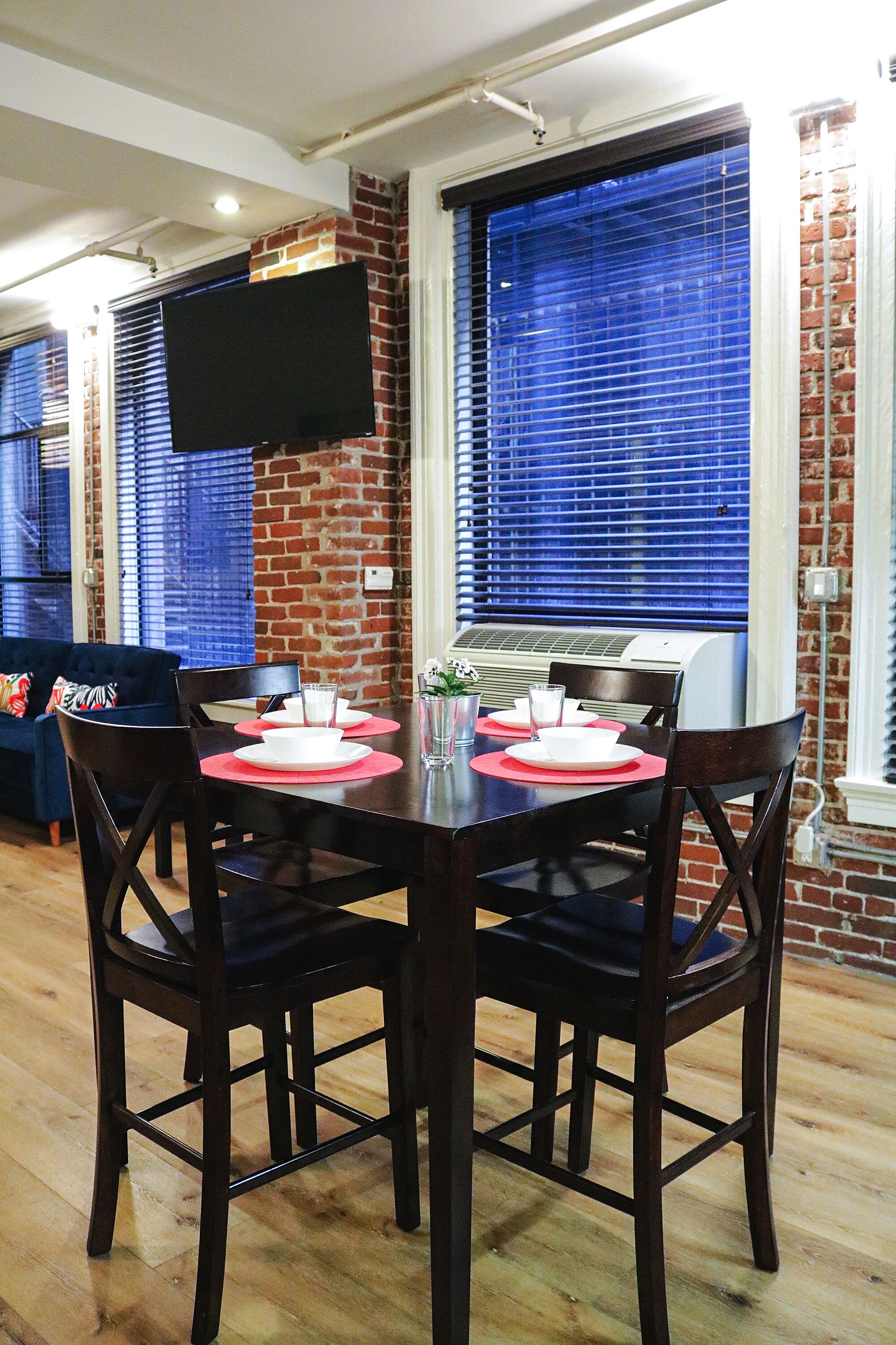 A dining area with a brick wall, large window with blinds, a flat-screen TV mounted above the table, and a ceiling-mounted air conditioning unit.
