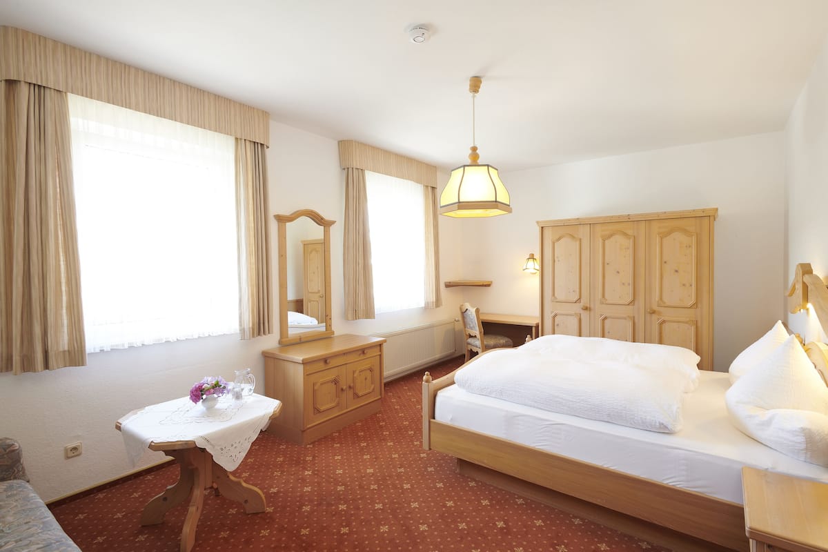 Habitación de hotel con cama, tocador, espejo, mesa con un florero y ventanas amplias con cortinas.