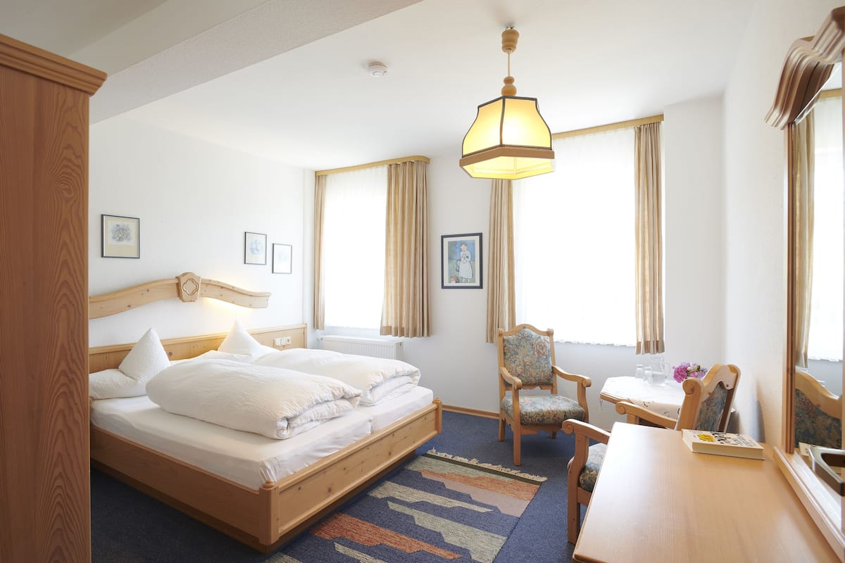 Habitación de hotel con una cama de madera, una mesita, una silla y un ventanal con cortinas.