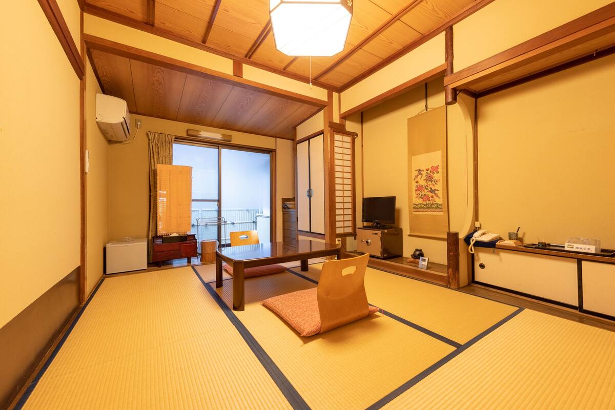 A traditional Japanese room with tatami flooring, a wooden table, a television, and a sliding door leading to a balcony.