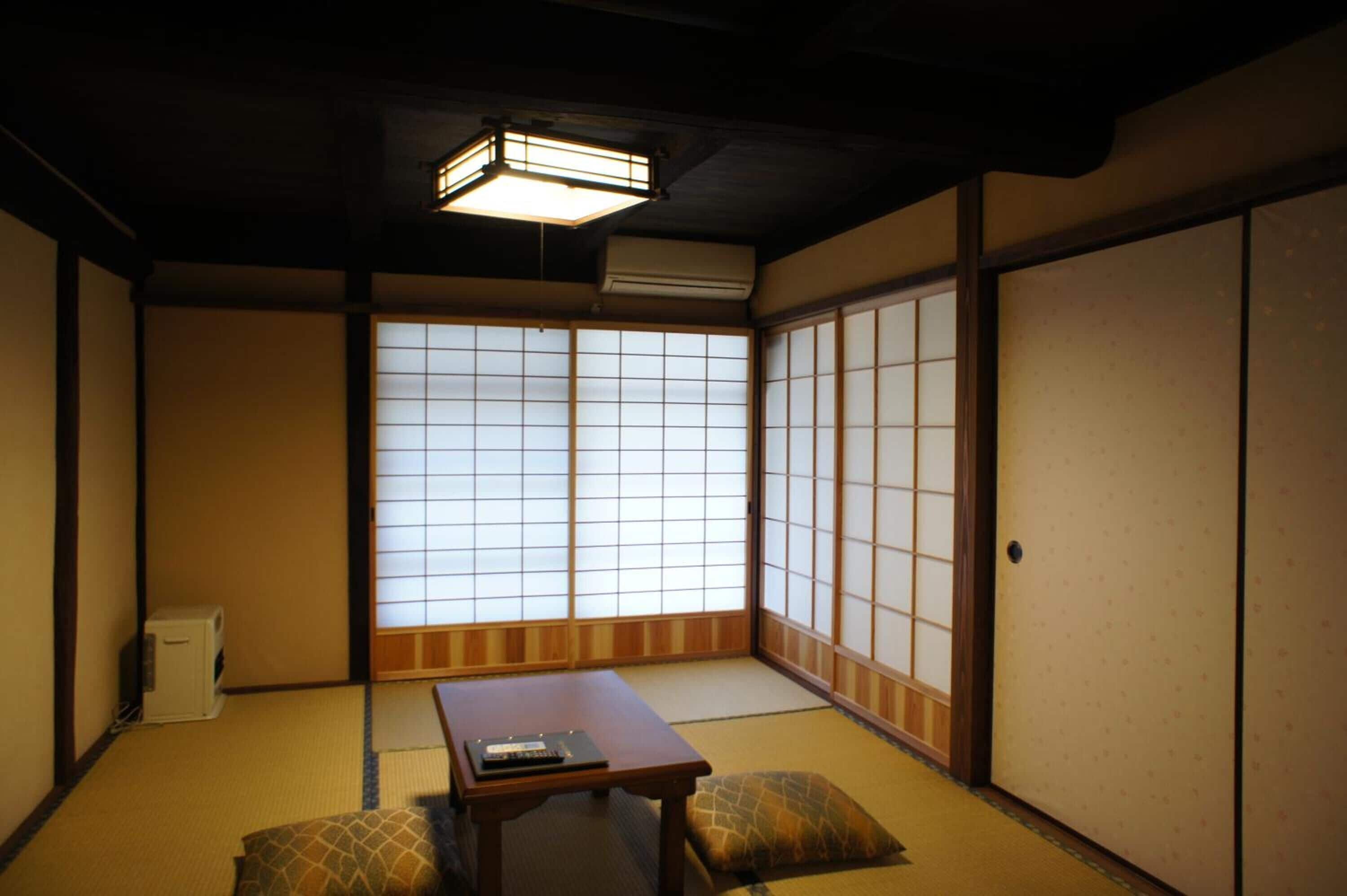 Ein traditioneller japanischer Raum mit Tatami-Böden, einem niedrigen Tisch, Kissen, verschiebbaren Shoji-Türen und einer Deckenleuchte.