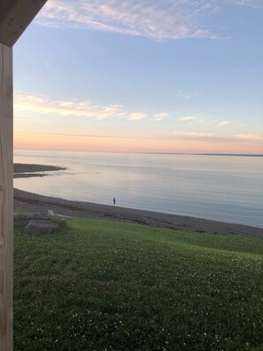 Blick auf einen Strand mit einer Person, die am Ufer entlanggeht, einer grünen Fläche im Vordergrund und einem ruhigen Meer unter blauem Himmel.