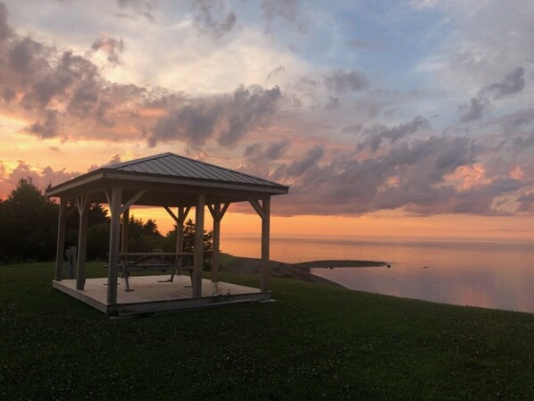 Ein Kiosk mit Metallveranda und offenen Seiten, auf einer Rasenfläche in der Nähe eines stillen Gewässers, unter einem Abendhimmel.