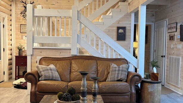 A spacious loft-style living area with a high wooden ceiling, a brown leather sofa, a wooden coffee table, and a staircase with white railings.