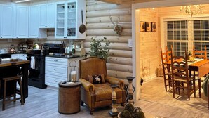 A spacious living area with a high ceiling, wooden beams, a dining table with chairs, a cozy armchair, and a kitchen in the background.