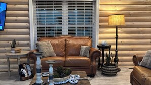 A cozy log cabin interior with a leather sofa, a wooden coffee table, and a large window with blinds.