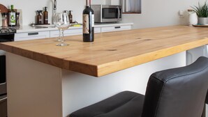 Una cocina con una isla de madera, una botella de vino y dos copas. Hay un microondas, un fregadero y una estantería con cuencos y tazas.