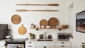 Una cocina con una mesa de madera, armarios blancos, una estufa, un microondas y un estante con vajilla y botellas.