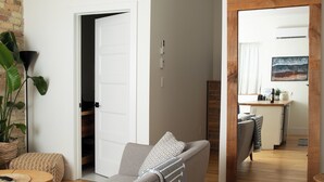 Una sala de estar moderna con un sofá gris, una mesa de centro y una planta grande. Se ve un suelo de madera y una zona de cocina a través de una puerta abierta.