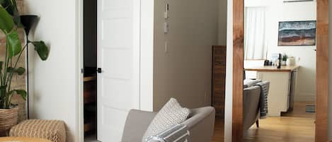 Una sala de estar moderna con un sofá gris, una mesa de centro y una planta grande. Se ve un suelo de madera y una zona de cocina a través de una puerta abierta.
