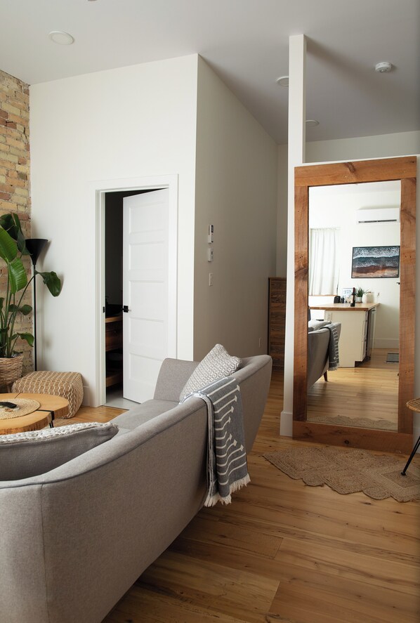 A modern living room with a grey sofa, a coffee table, and a large plant. A wooden floor and a kitchen area are visible through an open door.