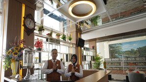A hotel reception area with a wooden reception desk, a clock, and a large window with a view of a brick wall and greenery.
