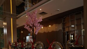 A modern hotel lobby with a long reception counter, decorative glassware, and a large potted plant with pink flowers.