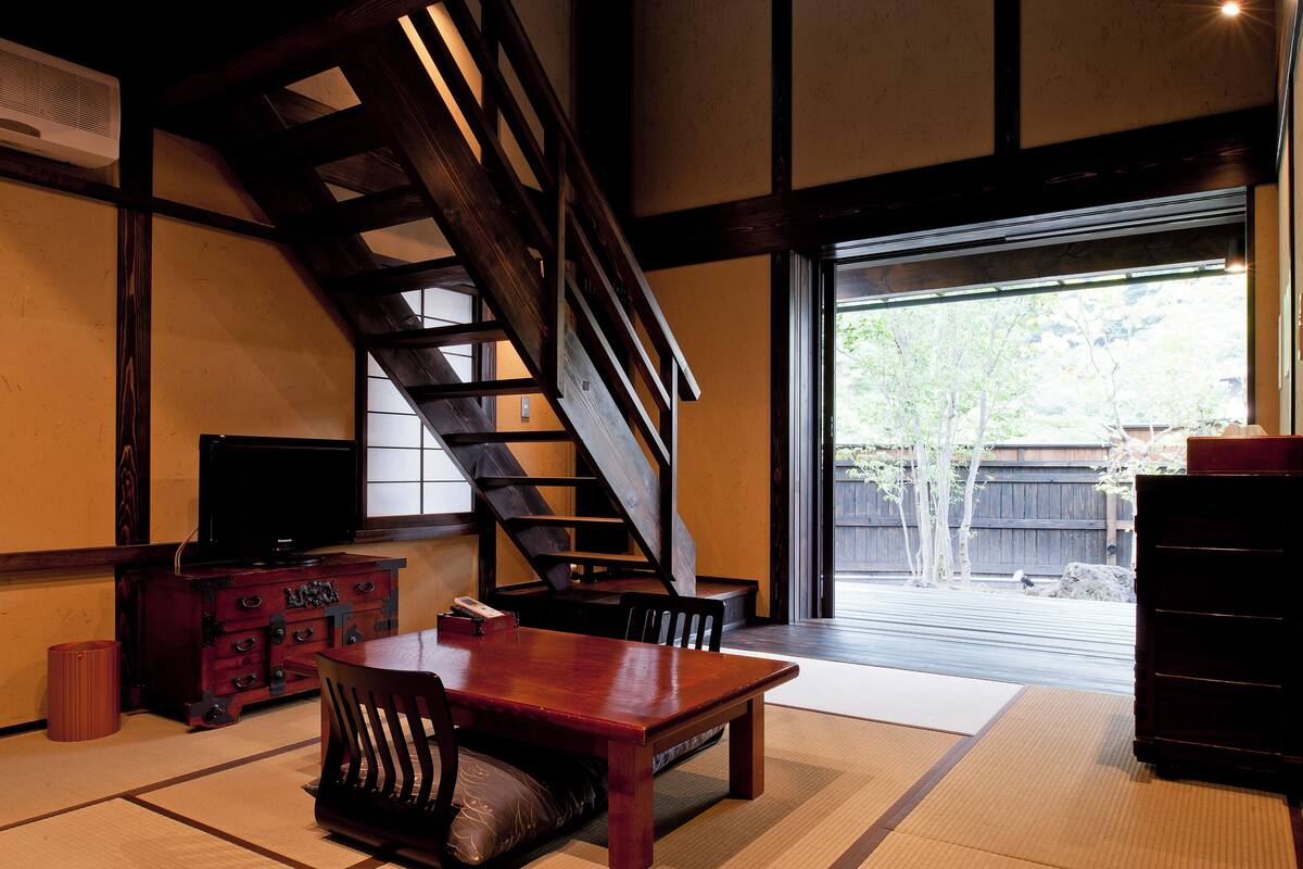 A traditional Japanese interior with tatami flooring, a wooden dining table, chairs, a television on a cabinet, and a sliding door leading to an outdoor area.