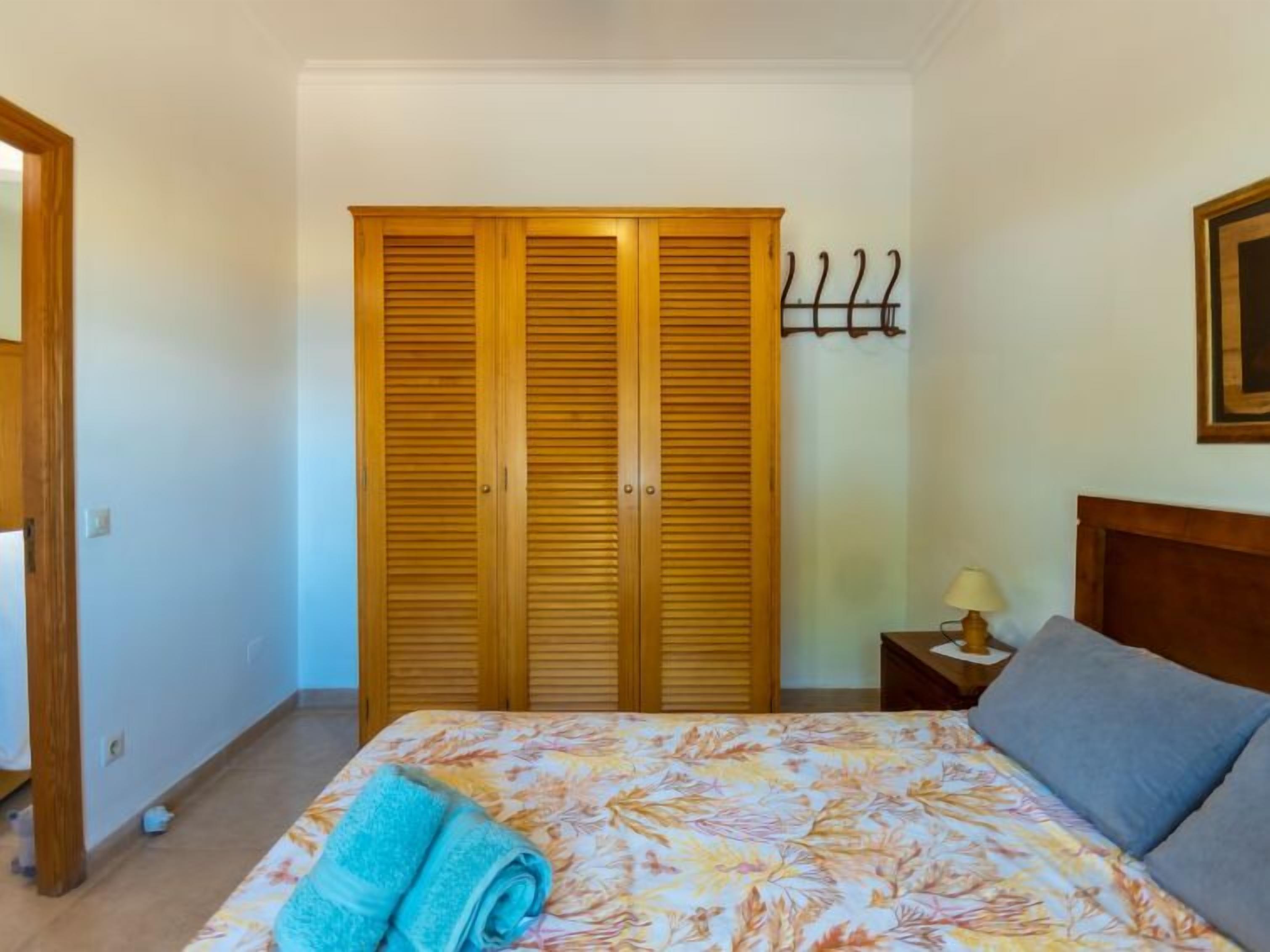 A bedroom with a bed, a wooden wardrobe, a nightstand with a lamp, and a coat rack.