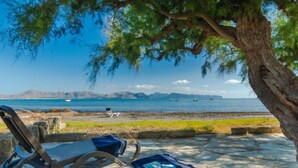 Ein Sitzbereich direkt am Strand mit Liegestühlen und einem Tisch, mit Blick auf ein ruhiges Meer und entfernte Berge.