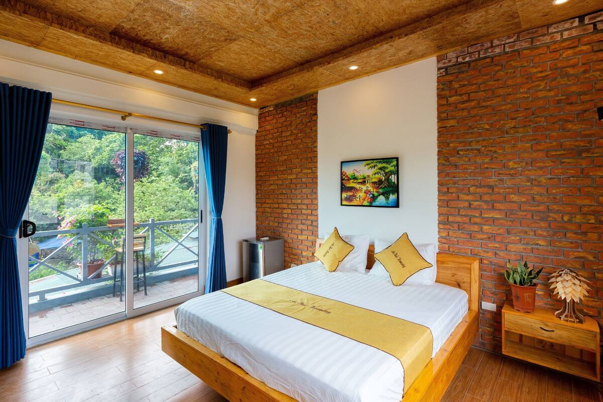 Une chambre avec un grand lit, un mur en briques, une fenêtre avec des rideaux et une petite table sur laquelle est posée une plante.