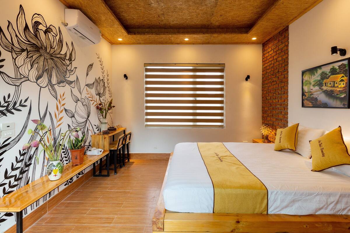Une chambre avec un grand lit, une tête de lit en bois, un bureau avec une chaise, une fresque murale, une fenêtre avec des stores et un mur en briques.