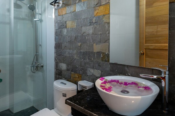 A bathroom with a stone wall, a white sink with flower petals, a shower, and a wooden door. - An Thai Bar & Homestay (Hoa Lu)
