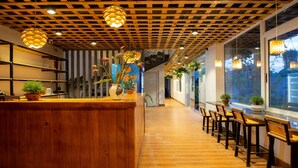 A wooden bar area with a long counter and multiple chairs, surrounded by large windows and potted plants. - An Thai Bar & Homestay (Hoa Lu)