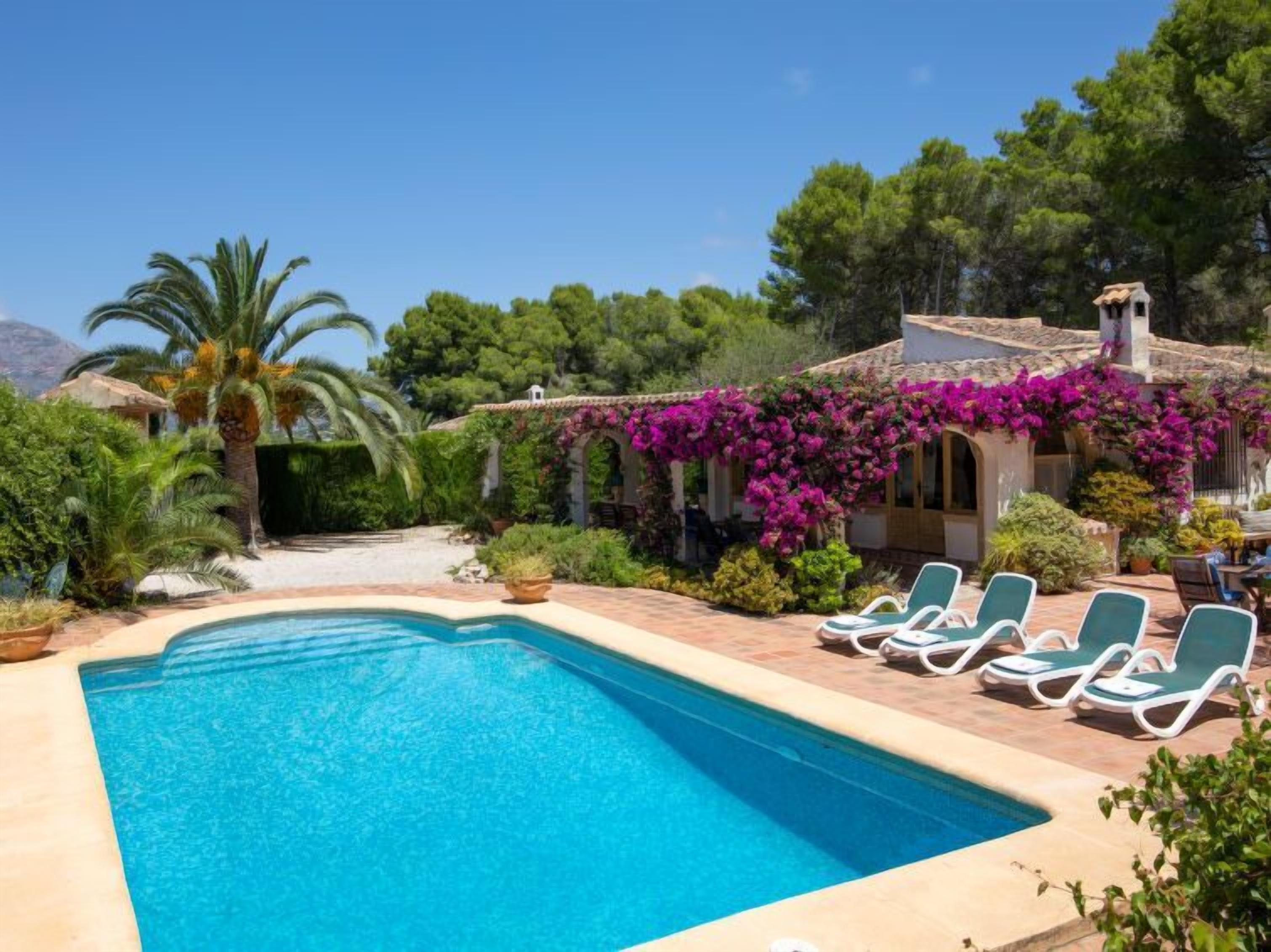 A swimming pool with lounge chairs, surrounded by lush greenery and a building with a tiled roof.