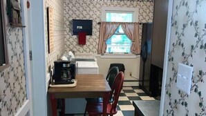 Una cocina con suelo a cuadros, una mesa de madera y sillas rojas. Se observa una ventana con cortinas y un refrigerador.