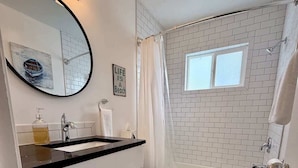 Ein Badezimmer mit weißem Waschbecken, Spiegel und Toilette, einer Duschkabine mit Vorhang sowie einem Fenster.