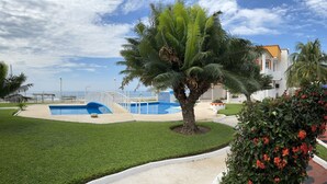 A pool area with a blue pool, palm trees, and a red car parked near a building with a white exterior. - Villas del Pacifico | Habitación Privada #4  (El Carrizal)