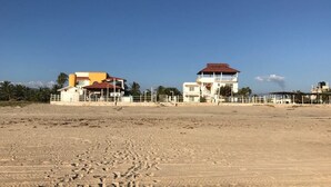 A beach with footprints leading to a white building with a red roof, surrounded by palm trees and a clear sky. - Villas del Pacifico | Habitación Privada #4  (El Carrizal)