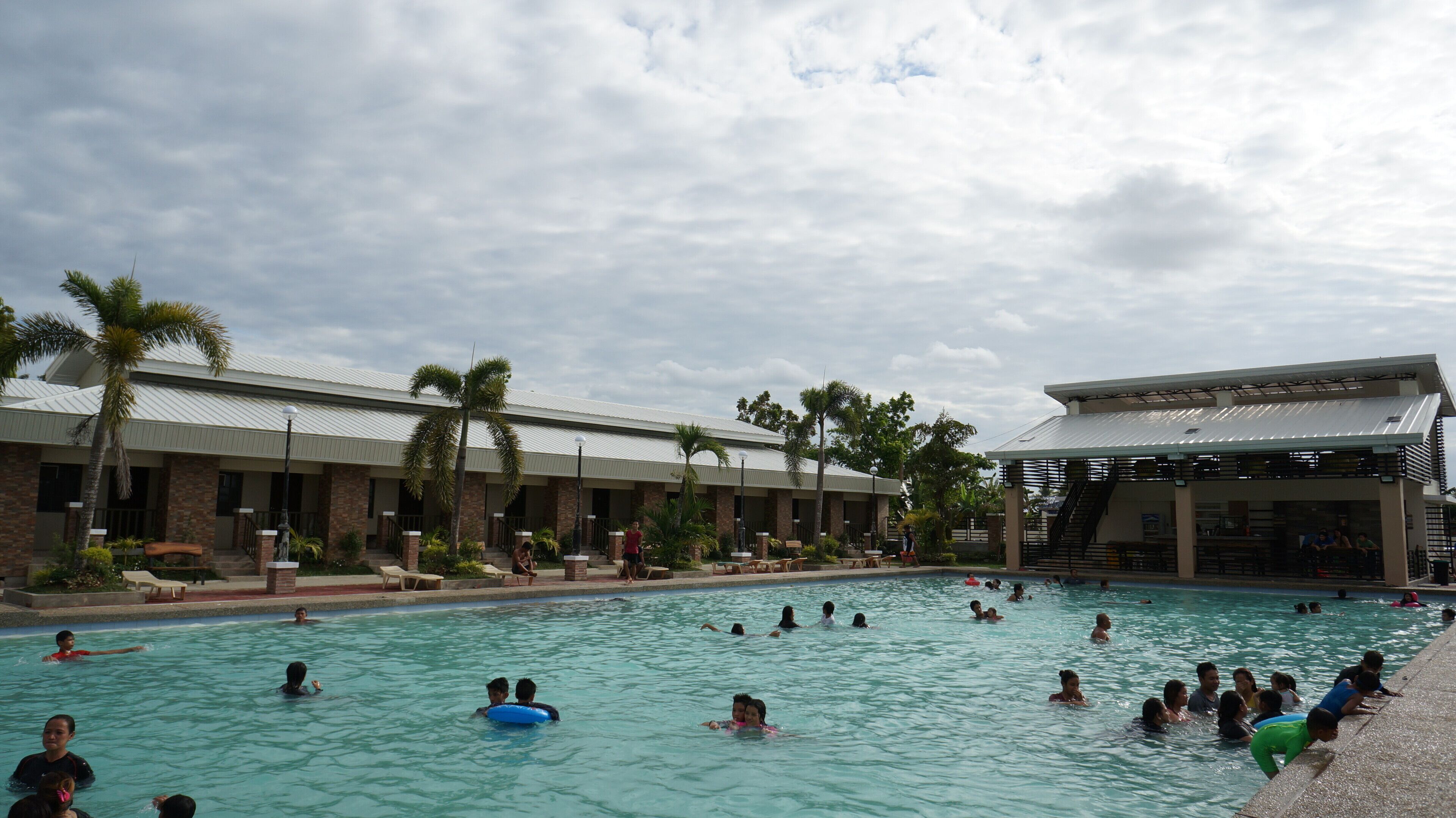 A swimming pool area with multiple people swimming and lounging, surrounded by palm trees and a building with a covered seating area.