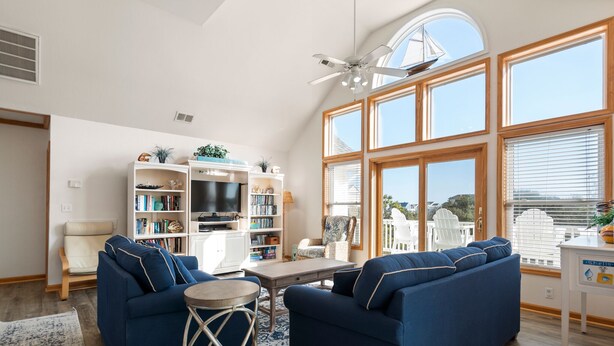 A spacious living room with a high ceiling, large windows, a ceiling fan, a fireplace, a bookshelf, a TV, and a seating area with a sofa and chairs. - Shell's Shack: Oceanside, Private Pool, Hot Tub, Tiki Bar, Putting Green (Corolla)