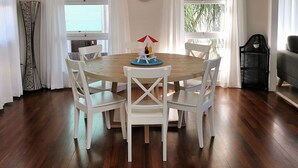 A dining area with a round wooden table surrounded by white chairs, a red and white umbrella, and a small table with condiments. - Beach Shack On 115 Esplanade (BARGARA)