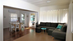 A living room with a dark grey sofa, a small round table, and a dining area with white chairs. - Beach Shack On 115 Esplanade (BARGARA)