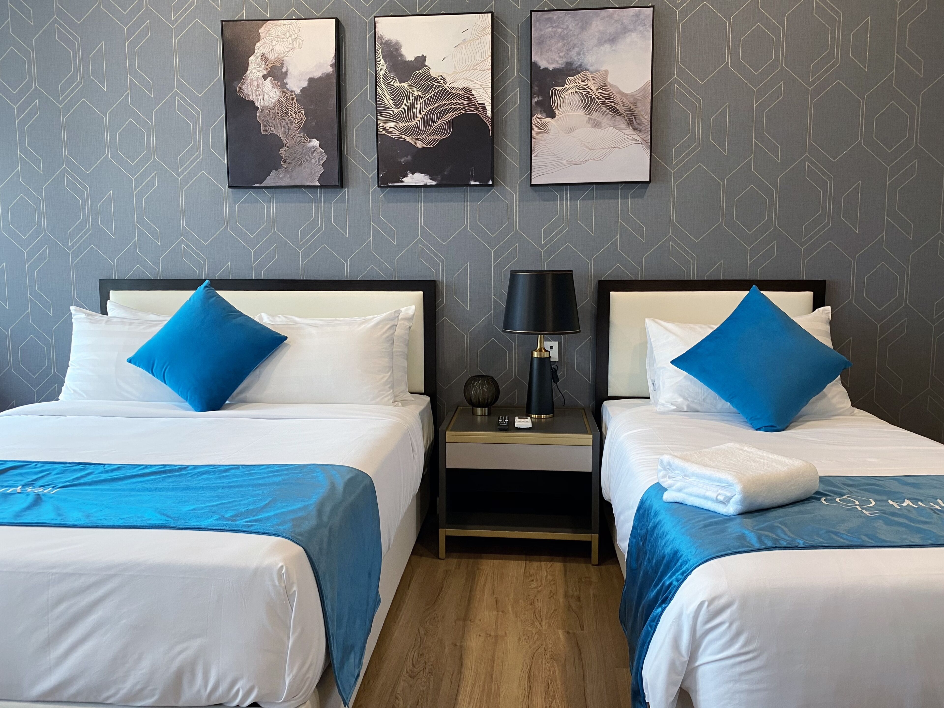 A hotel room with two beds, each with a blue bedspread and white pillows, a nightstand with a lamp, and three framed artworks on the wall.