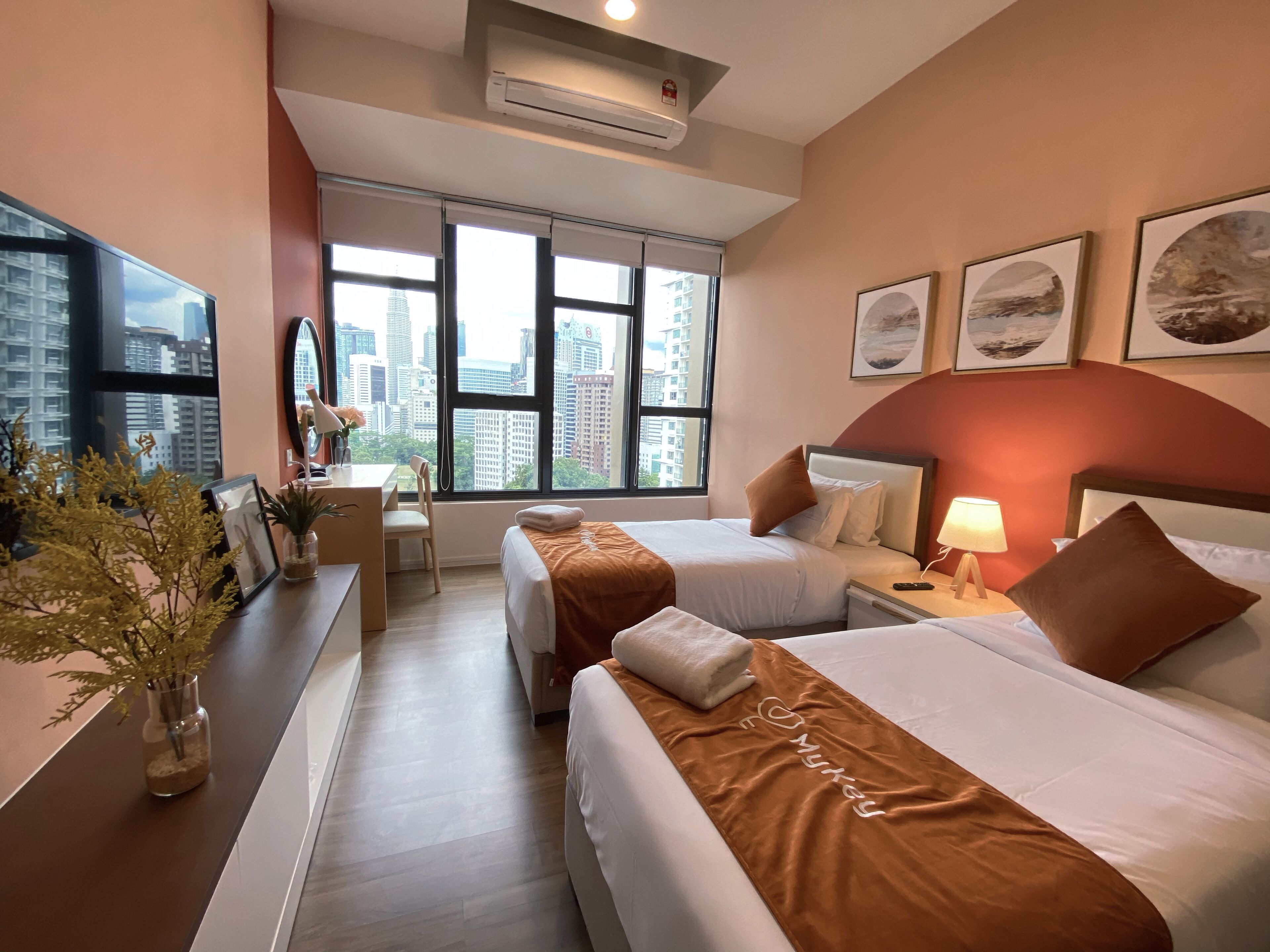 Habitación de hotel con dos camas, un escritorio y una silla. Amplias ventanas con vista a la ciudad, un televisor y obras de arte enmarcadas en la pared.