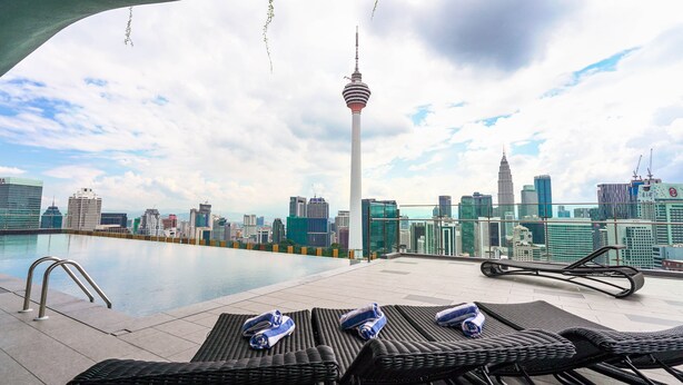 Zona de piscina en la azotea con sillones de descanso y toallas, con vista a un horizonte urbano de edificios modernos y una torre destacada.