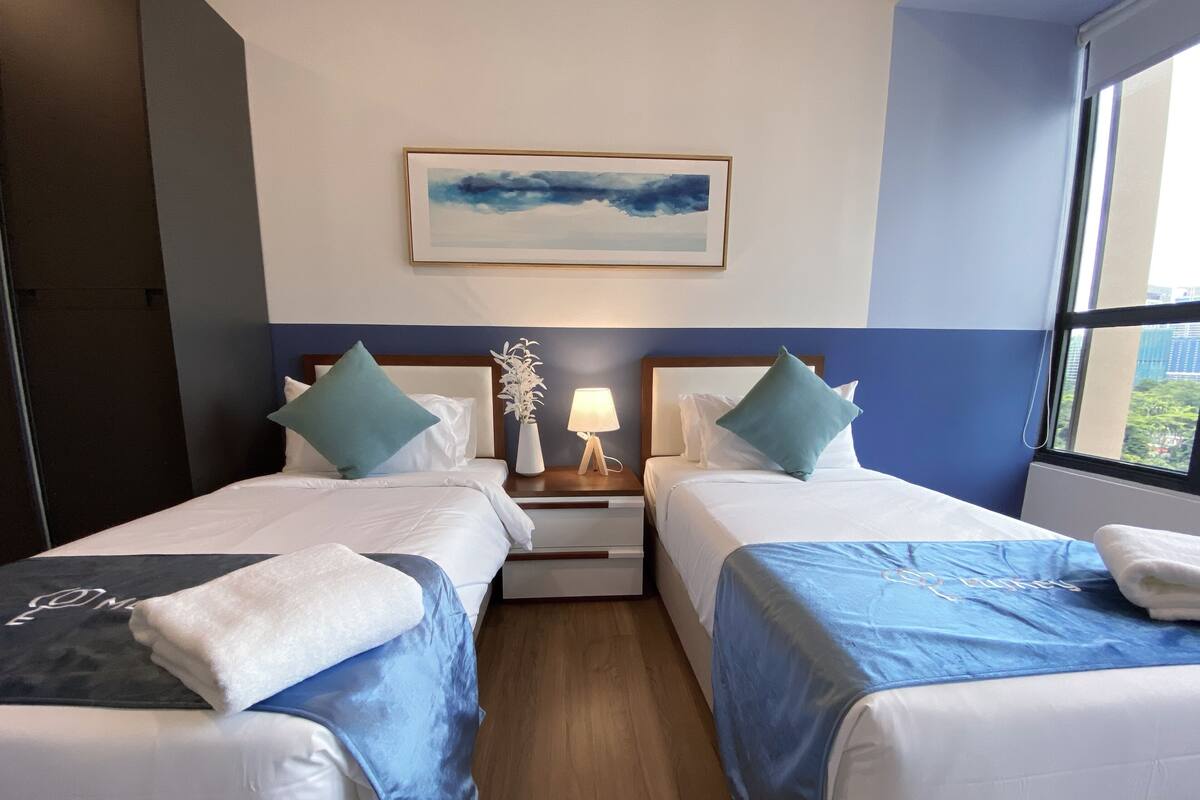 A hotel room with two beds, a nightstand with a lamp, a framed picture on the wall, and a window with a view.
