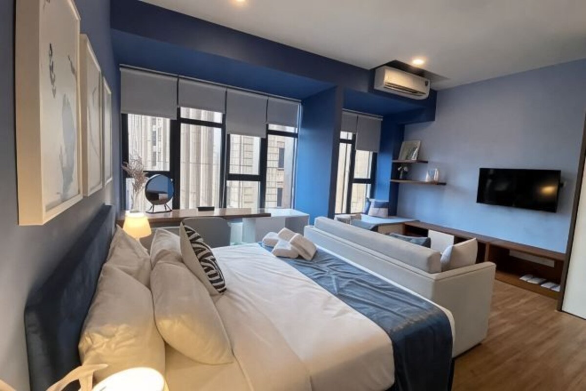 A modern hotel room with a bed, sofa, TV, and large windows.