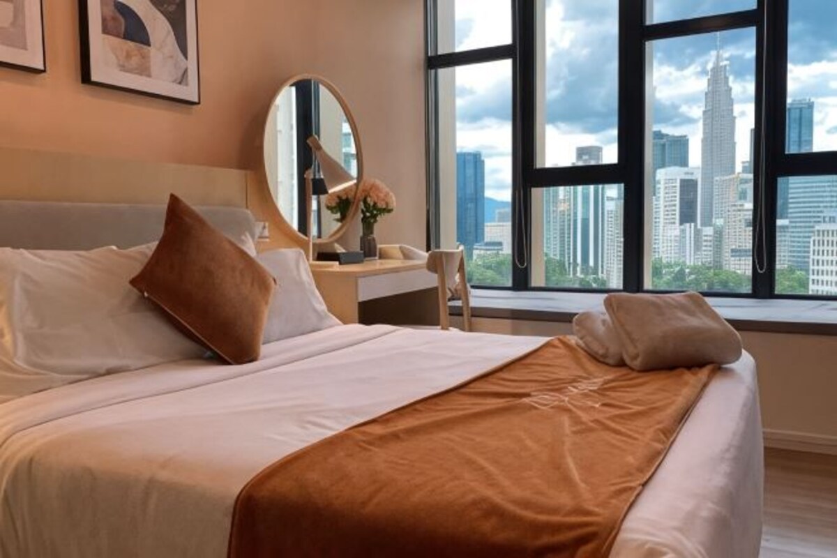 A hotel room with a bed, a desk, a chair, a mirror, and a view of a cityscape through large windows.