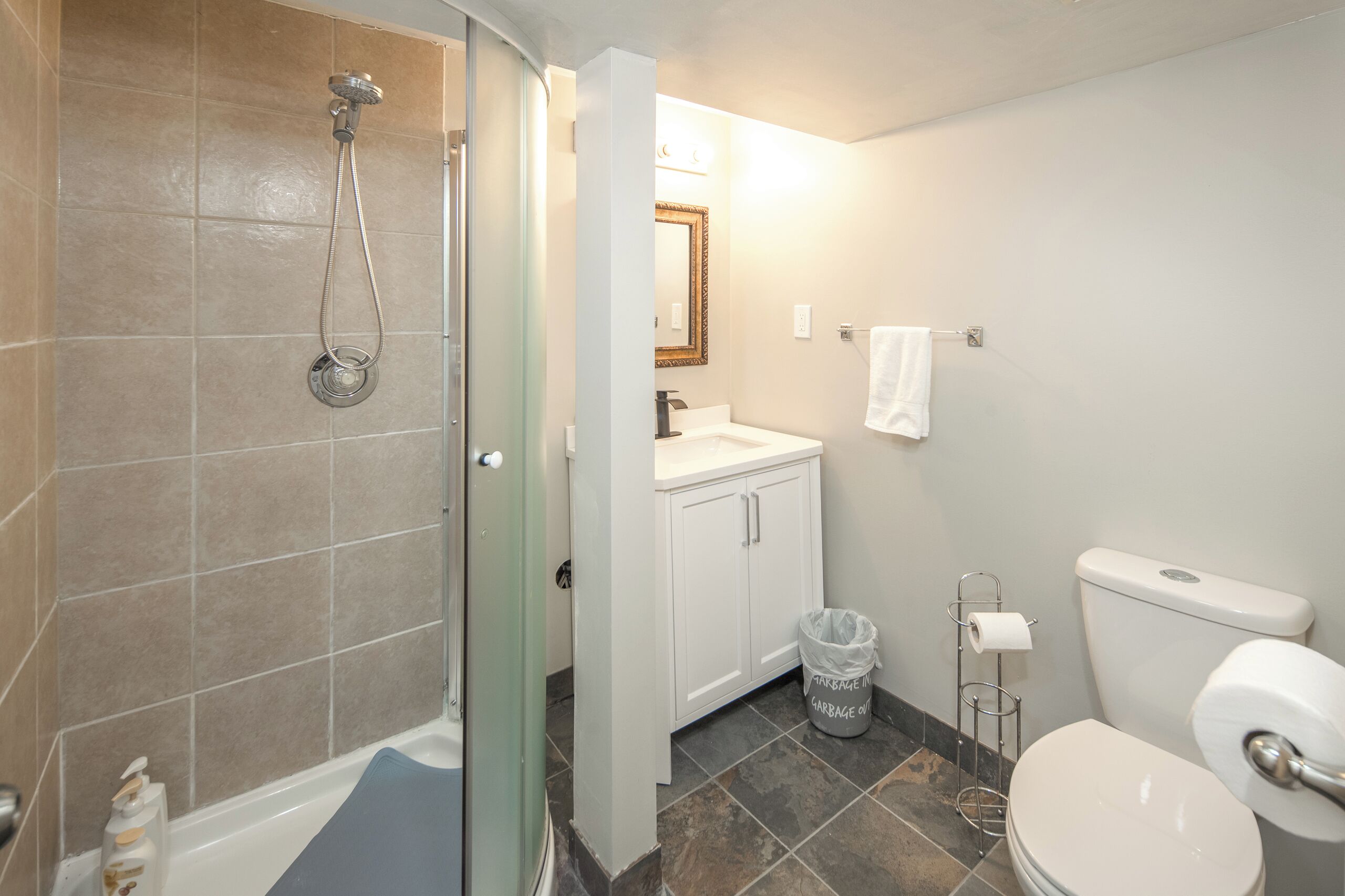 A bathroom with a shower, toilet, sink, and a trash can.