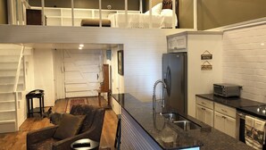 Una cocina moderna con una isla integrada, un refrigerador y un tostador. Hay una escalera que conduce a un dormitorio tipo loft con una cama y una silla.