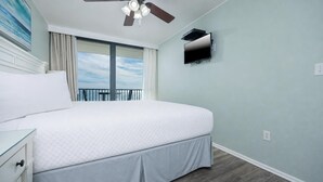 A bedroom with a bed, a ceiling fan, a TV mounted on the wall, and a view of the sky through the window.