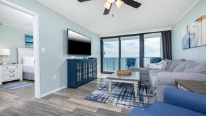 A modern living room with a flat-screen TV, a sofa, a coffee table, and a view of the ocean through large windows.