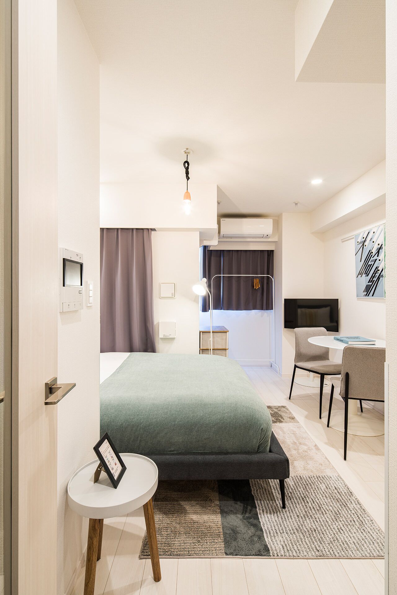 A modern hotel room with a bed, bedside table, desk, chair, and an air conditioning unit.