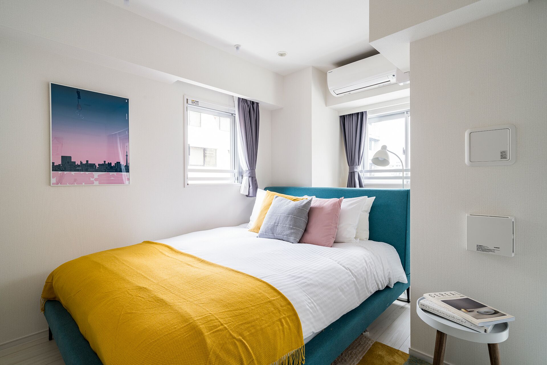 A modern bedroom with a teal headboard, a yellow throw blanket, and a cityscape painting on the wall.