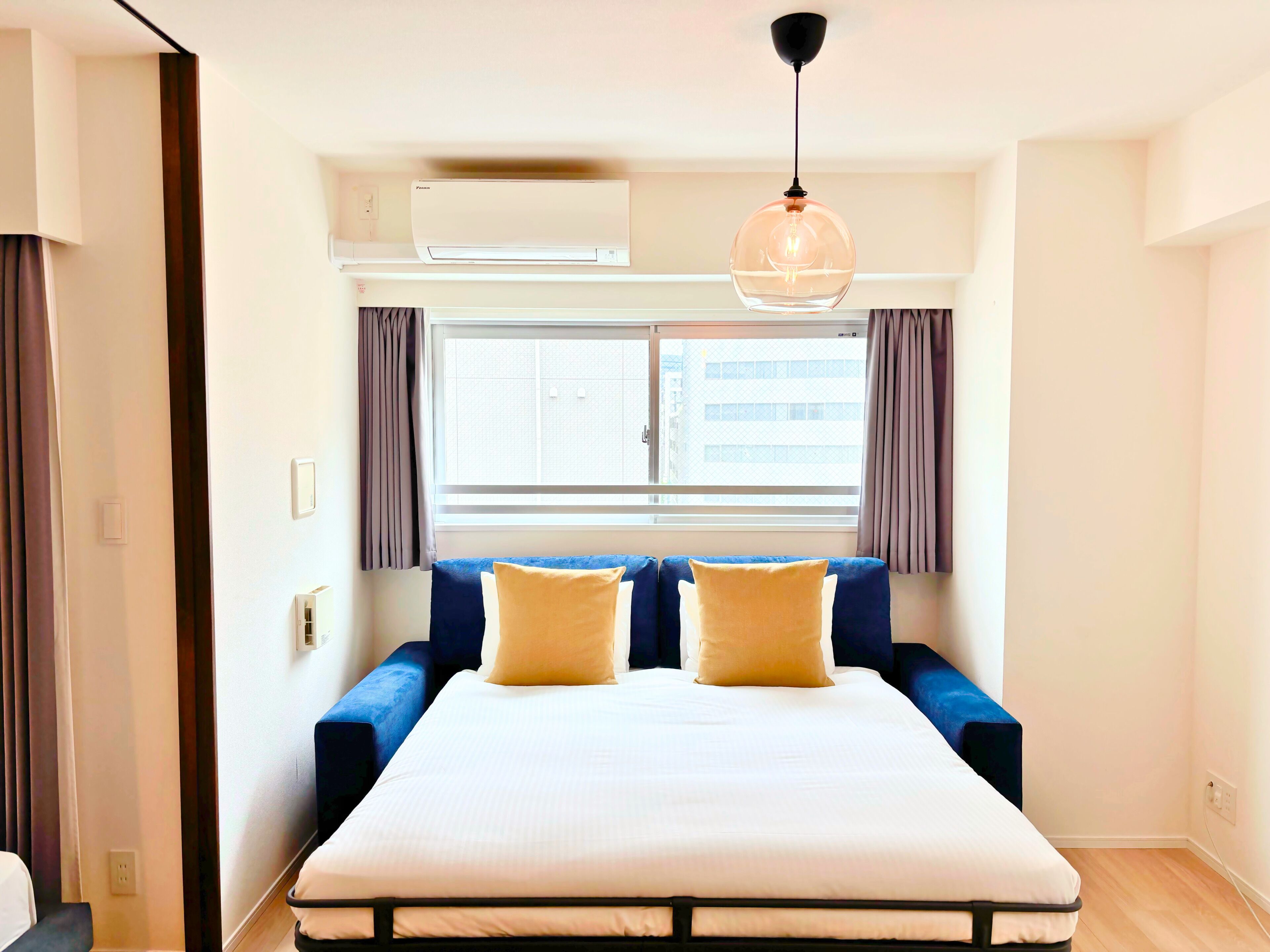 A modern hotel room with a large bed, a sofa, a window with curtains, an air conditioning unit, and a hanging light fixture.