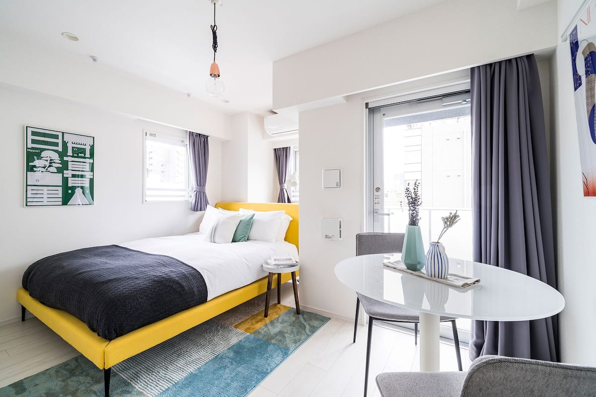 Quarto de hotel moderno com uma cama, uma mesa pequena e uma cadeira. Há uma janela com cortinas, um ar condicionado fixado na parede e um quadro na parede.